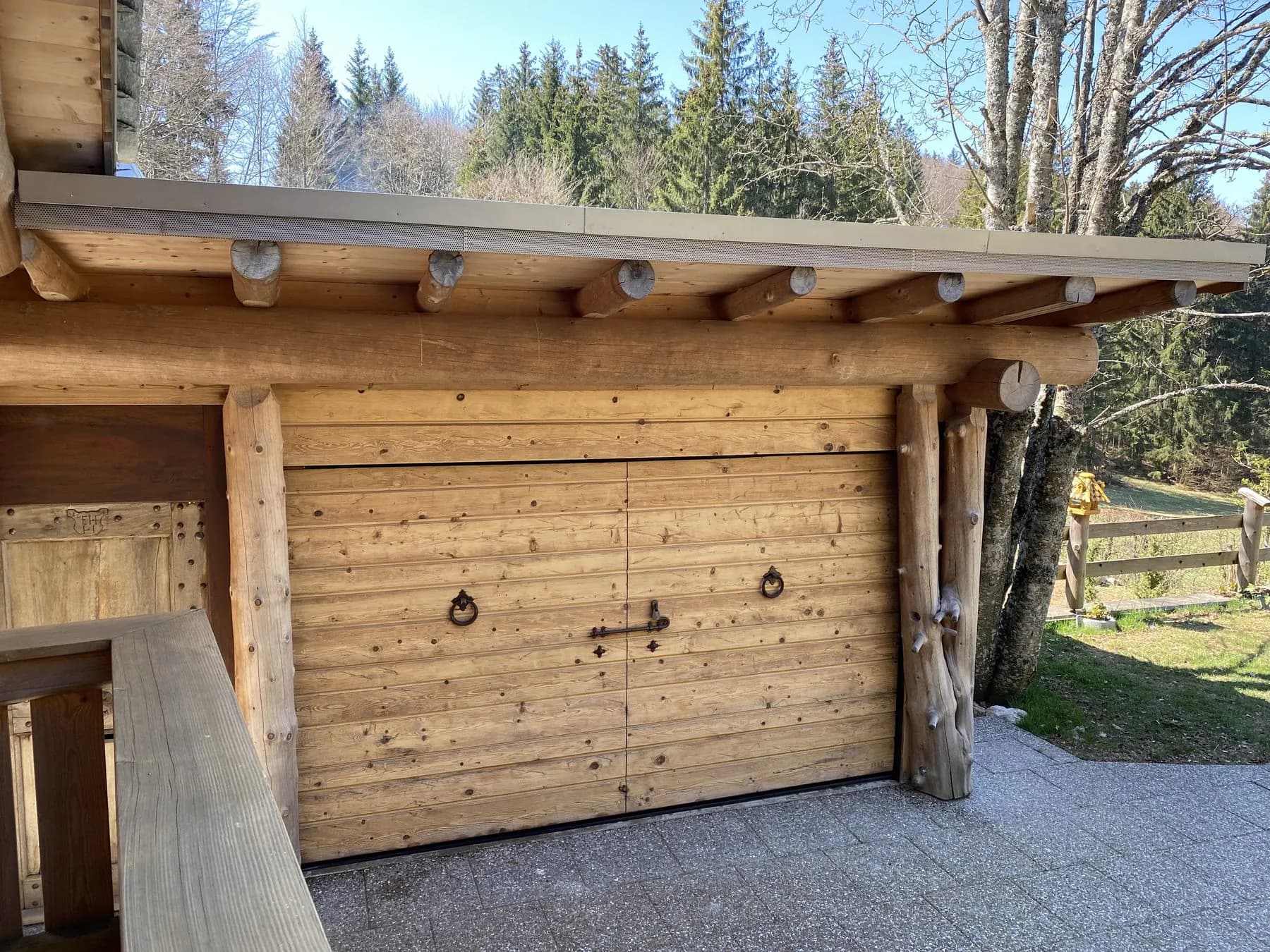 Echtholztor mit geschmiedeten Ringgriffen an einem Bau aus Rundstämmen, dahinter Wald und Wiese