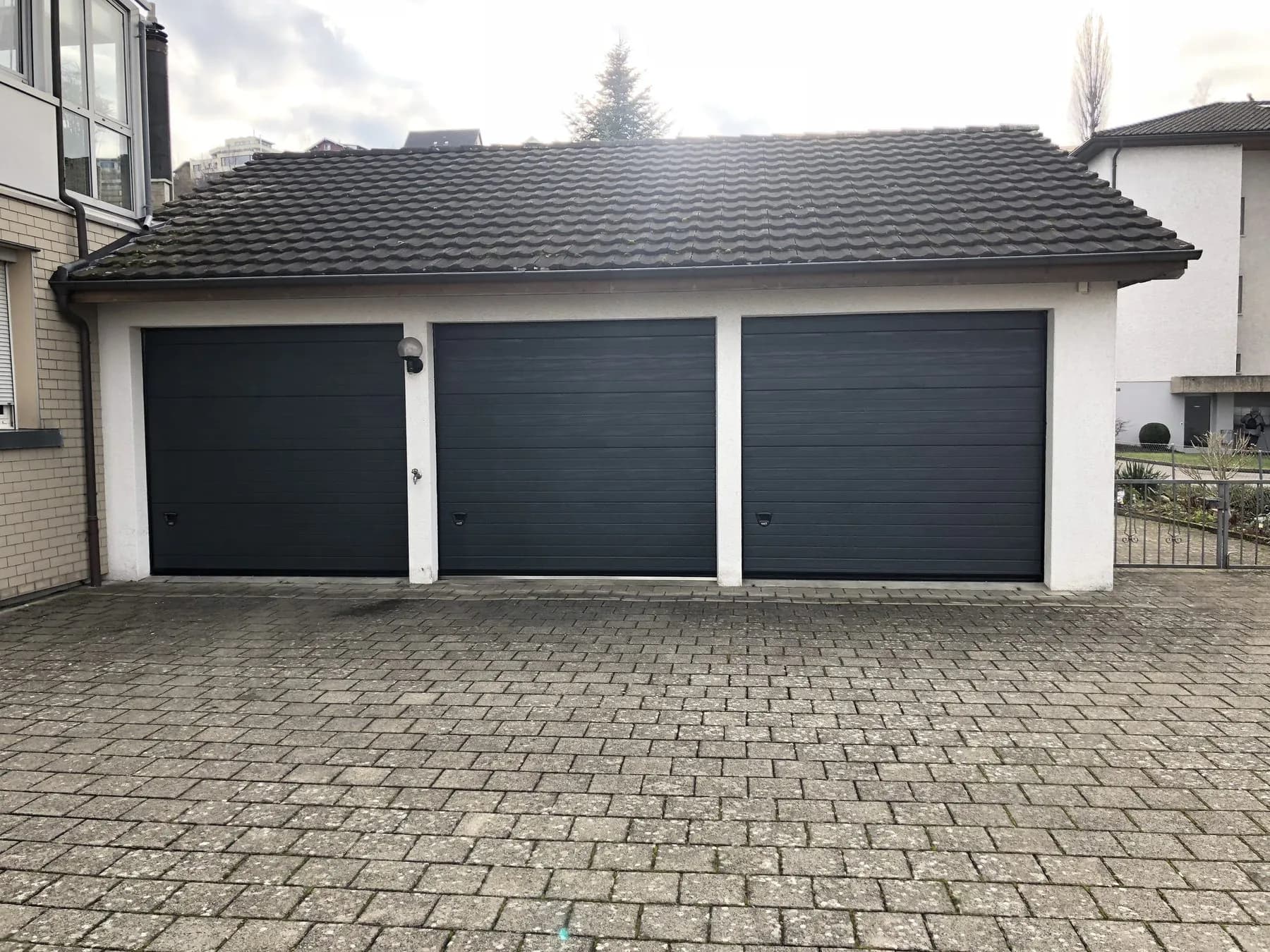 Drei anthrazitfarbene Sektionaltore an einem Garagenbau mit Ziegeldach und gepflastertem Vorplatz