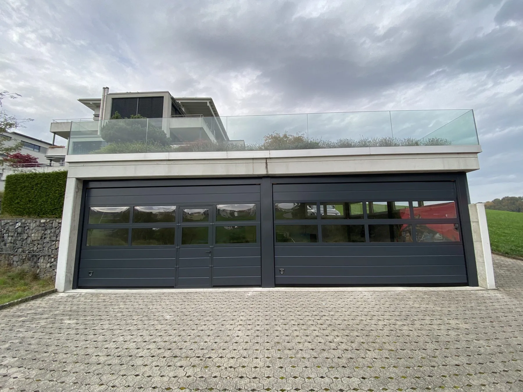 Zwei anthrazitfarbene Sektionaltore mit Lichtband und eingebauter Tür unter der Terrasse eines Wohnhauses