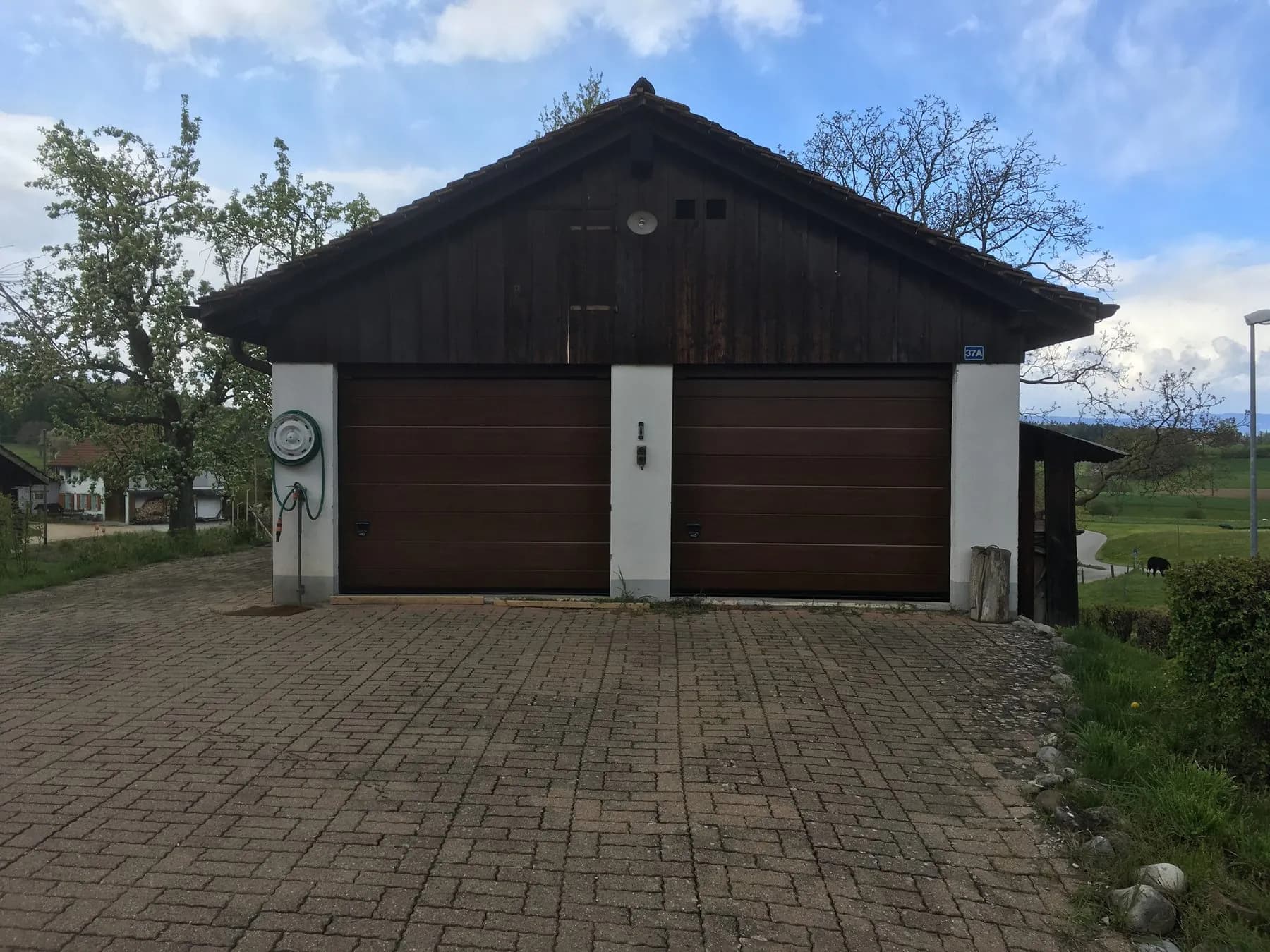 Zwei braune Sektionaltore in einem Ökonomiegebäude mit dunkler Holzverschalung im Giebel, ringsum Wiesen und Obstbäume
